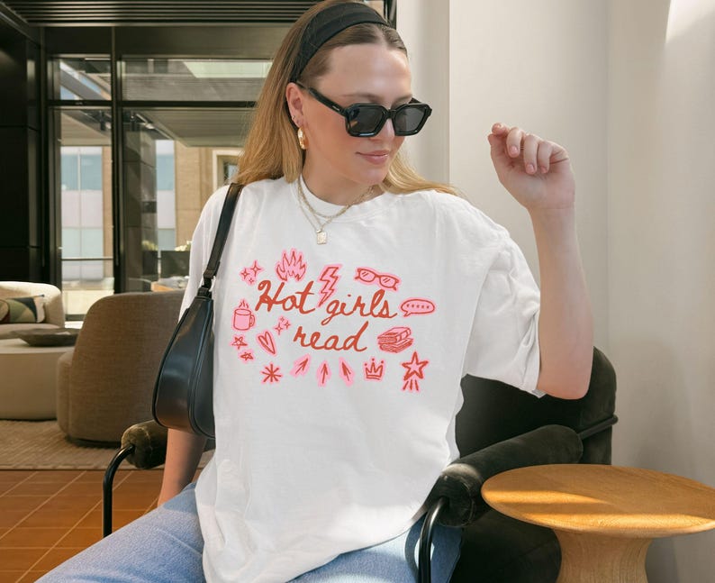 May include: White t-shirt with the text "Hot girls read" in pink, along with various pink graphic elements. The shirt is worn by a person wearing black sunglasses and a black headband.
