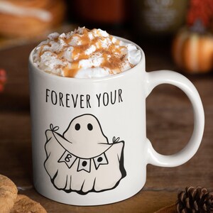 May include: White ceramic mug with the text "FOREVER YOUR" and a ghost illustration. The mug is filled with a beverage topped with whipped cream and a sprinkle of cinnamon. A festive Halloween-themed mug.