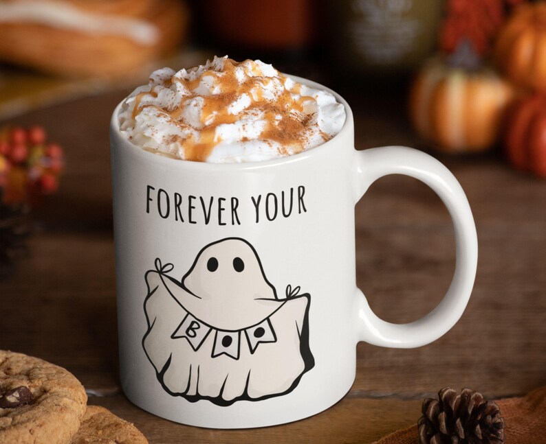 May include: White ceramic mug with the text "FOREVER YOUR" and a ghost illustration. The mug is filled with a beverage topped with whipped cream and a sprinkle of cinnamon. A festive Halloween-themed mug.