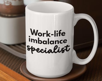 May include: White ceramic mug with the text "Work-life imbalance Specialist" in black. The mug has a large handle and sits on a brown saucer. The mug is a humorous gift idea for a coworker or friend.