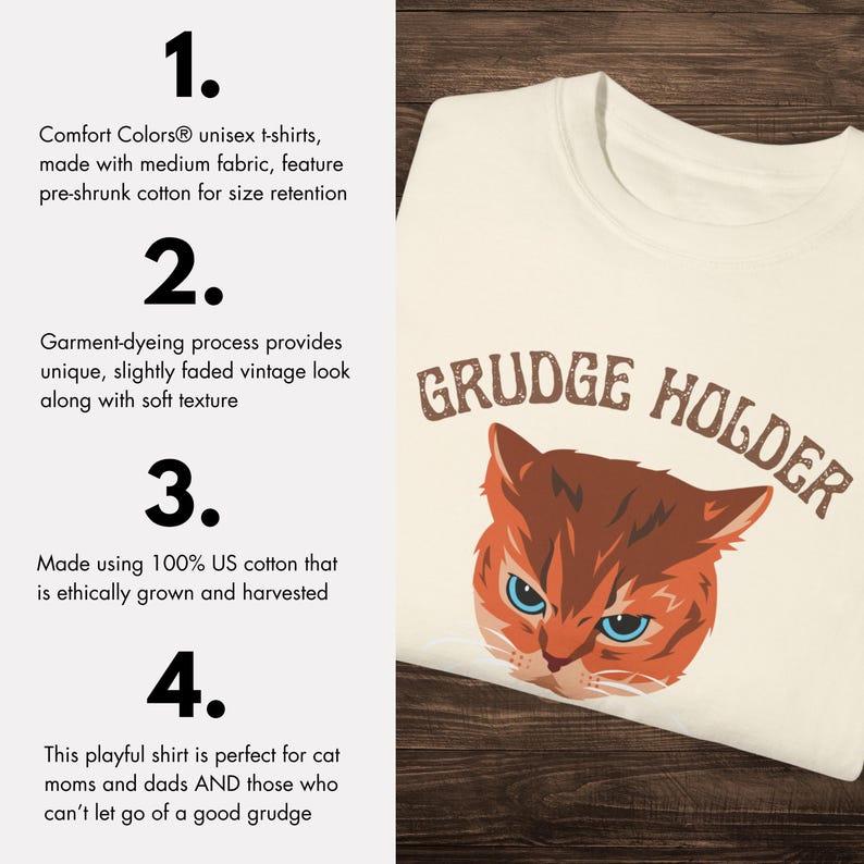 May include: White t-shirt with a cartoon cat face and the text "GRUDGE HOLDER" in brown lettering. The shirt is made of 100% US cotton.
