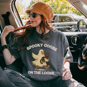 May include: Dark gray t-shirt with the text "Spooky Goose On The Loose" and a cartoon goose dressed as a ghost, holding a black cat. The shirt is worn by a person in a car, wearing sunglasses and a brown hat.