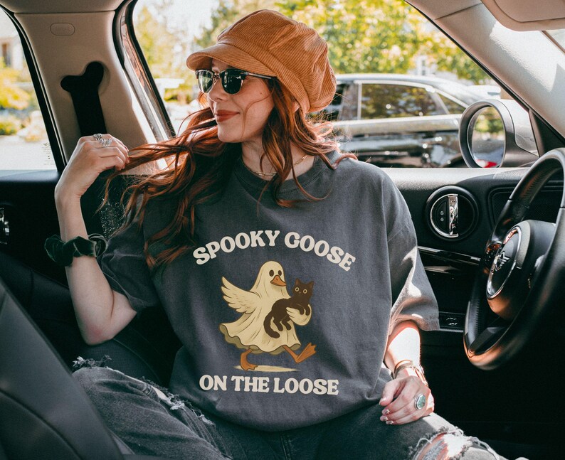 May include: Dark gray t-shirt with the text "Spooky Goose On The Loose" and a cartoon goose dressed as a ghost, holding a black cat. The shirt is worn by a person in a car, wearing sunglasses and a brown hat.