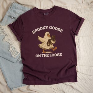 May include: A maroon t-shirt with the text "Spooky Goose on the Loose" in white. The graphic features a goose in a ghost costume holding a black cat. Light blue denim shorts are visible in the background.
