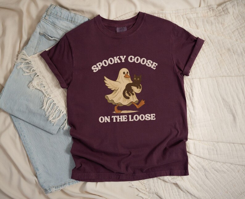 May include: A maroon t-shirt with the text "Spooky Goose on the Loose" in white. The graphic features a goose in a ghost costume holding a black cat. Light blue denim shorts are visible in the background.