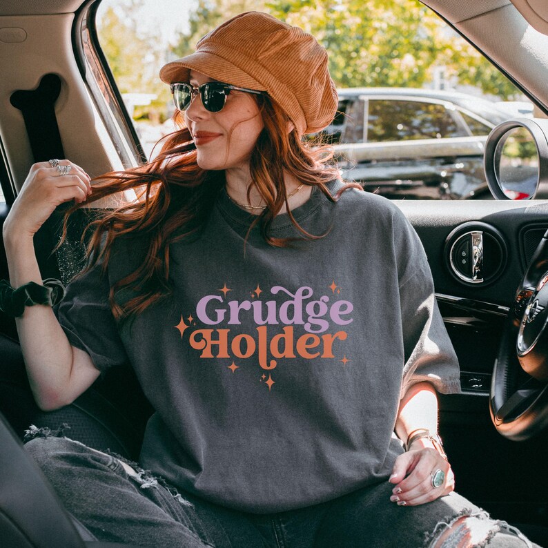 May include: A woman wearing a gray t-shirt with the text "Grudge Holder" in a retro font with pink and orange accents. The shirt has a distressed look and is decorated with small stars.