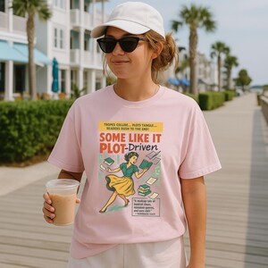 May include: Pale pink t-shirt with the text "SOME LIKE IT PLOT-DRIVEN" in red and green. The shirt features a vintage-style illustration of a woman surrounded by books. The text "TROPES COLLIDE... PLOTS TANGLE... READERS RUSH TO THE END!" is above the main text.