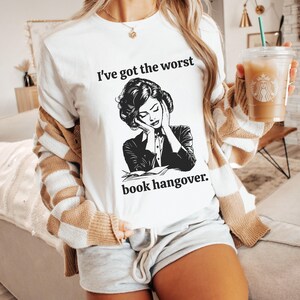 Op de afbeelding: Wit T-shirt met zwarte tekst die "Ik heb de ergste kater van een boek." luidt. Onder de tekst staat een zwart-witte illustratie van een vrouw met haar hoofd in haar handen.