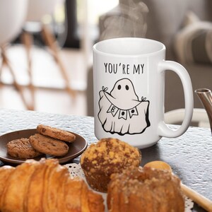 May include: White ceramic mug with the text "YOU'RE MY" and a cartoon ghost illustration. The mug is on a table with pastries and cookies. Steam rises from the mug, suggesting a hot beverage.