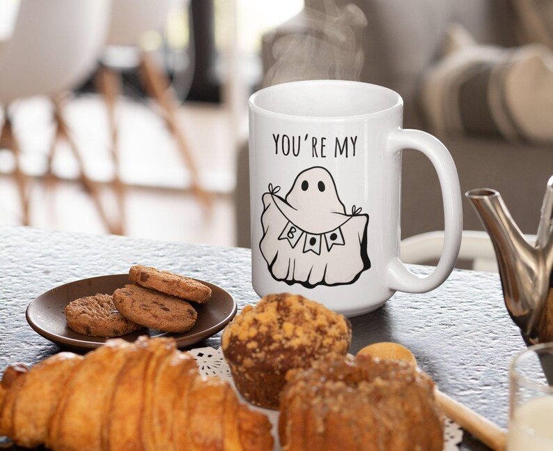 May include: White ceramic mug with the text "YOU'RE MY" and a cartoon ghost illustration. The mug is on a table with pastries and cookies. Steam rises from the mug, suggesting a hot beverage.