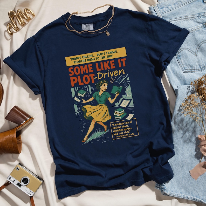 May include: Dark blue t-shirt featuring the text "SOME LIKE IT PLOT-Driven" and a retro illustration of a woman with books. The shirt also includes the text "TROPES COLLIDE... PLOTS TANGLE... READERS RUSH TO THE END!" and "A madcap tale of bookish chaos, mistaken genres, and zero chill."