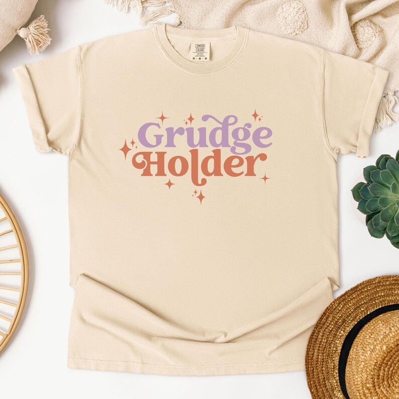 May include: A beige t-shirt with the text "Grudge Holder" in a retro font with purple and orange colors and small star accents.