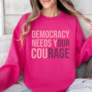 Puede incluir: Una sudadera rosa brillante con el texto "DEMOCRACY NEEDS YOUR COURAGE" en un dise&ntilde;o degradado en capas. La sudadera es de color liso y tiene mangas largas.