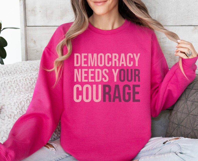 Puede incluir: Una sudadera rosa brillante con el texto "DEMOCRACY NEEDS YOUR COURAGE" en un dise&ntilde;o degradado en capas. La sudadera es de color liso y tiene mangas largas.