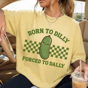 May include: Pale yellow t-shirt with a green cartoon pickle and the text "BORN TO DILLY FORCED TO DALLY". The design includes a green and white chequered pattern. The shirt is a light yellow colour.
