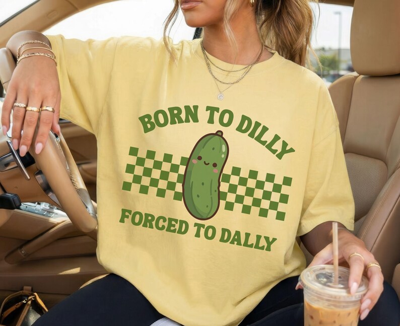 May include: Pale yellow t-shirt with a green cartoon pickle and the text "BORN TO DILLY FORCED TO DALLY". The design includes a green and white chequered pattern. The shirt is a light yellow colour.