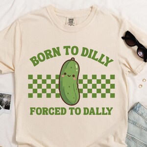 May include: A cream-coloured t-shirt with the text "BORN TO DILLY FORCED TO DALLY" in green. The design features a cartoon pickle with a smiling face, above a green and white checkered pattern. Sunglasses and denim shorts are also visible.