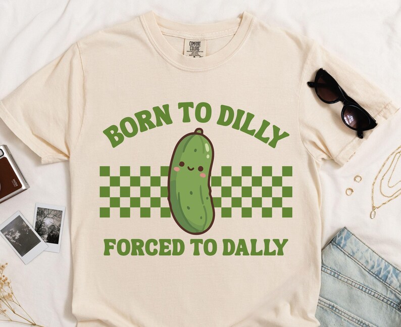 May include: A cream-coloured t-shirt with the text "BORN TO DILLY FORCED TO DALLY" in green. The design features a cartoon pickle with a smiling face, above a green and white checkered pattern. Sunglasses and denim shorts are also visible.