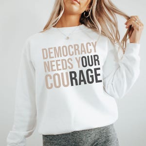 Puede incluir: Sudadera blanca con el texto "DEMOCRACY NEEDS YOUR COURAGE" en letras marrones y negras superpuestas. La sudadera es de cuello redondo y parece estar hecha de un material suave.