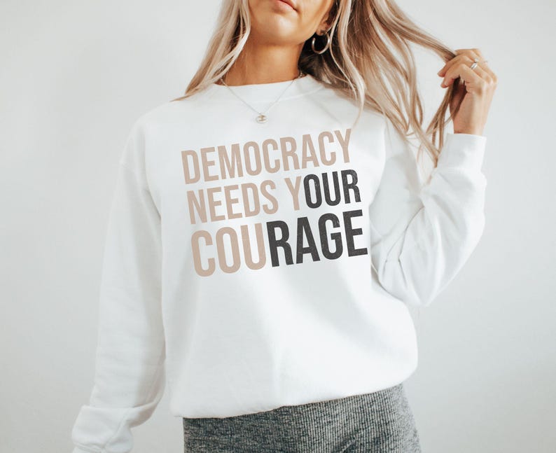 Puede incluir: Sudadera blanca con el texto "DEMOCRACY NEEDS YOUR COURAGE" en letras marrones y negras superpuestas. La sudadera es de cuello redondo y parece estar hecha de un material suave.