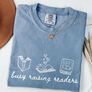 Może przedstawiać: Niebieski t-shirt z krótkim rękawem z białym nadrukiem graficznym przedstawiającym otwartą książkę, książkę z unoszącymi się sercami oraz książkę z ilustracją psa. Poniżej grafiki znajduje się biały napis "busy raising readers".