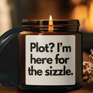Puede incluir: Una vela encendida en un tarro de vidrio marrón con tapa negra. La etiqueta dice "Plot? I'm here for the sizzle." El tarro está sobre una superficie de madera.