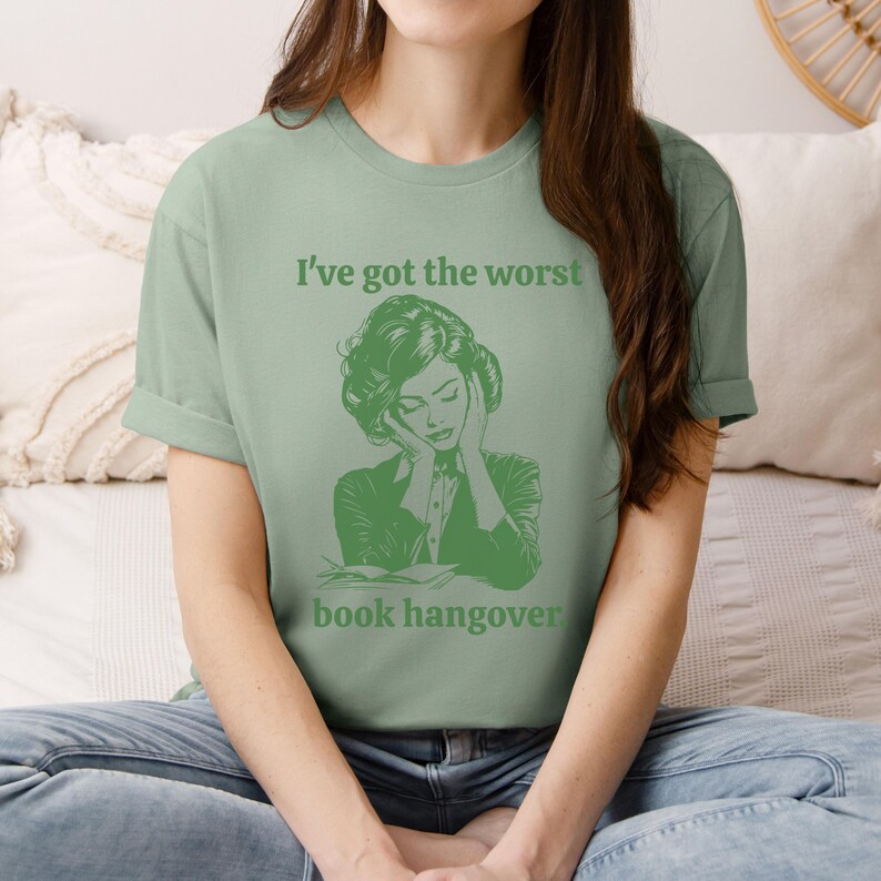 Op de afbeelding: Een groen T-shirt met de tekst "Ik heb de ergste kater van een boek" en een groene illustratie van een vrouw die haar hoofd in haar handen houdt.