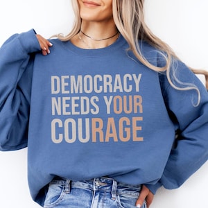 Puede incluir: Una sudadera azul con el texto "DEMOCRACY NEEDS YOUR COURAGE" en letras grises y melocot&oacute;n. La sudadera es de color liso y tiene mangas largas.