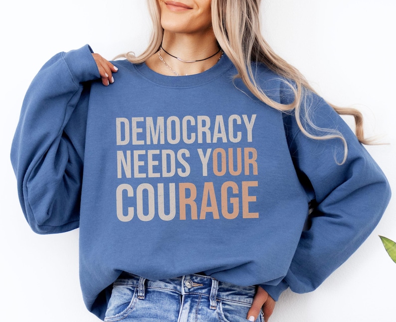 Puede incluir: Una sudadera azul con el texto "DEMOCRACY NEEDS YOUR COURAGE" en letras grises y melocot&oacute;n. La sudadera es de color liso y tiene mangas largas.