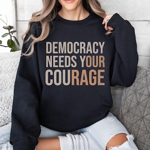 Puede incluir: Sudadera negra con el texto "DEMOCRACY NEEDS YOUR COURAGE" en una combinaci&oacute;n de colores neutros en capas. La sudadera es de cuello redondo y parece estar hecha de un material suave. El tema general es pol&iacute;tico e inspirador.