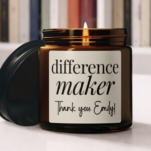 May include: A lit amber glass candle jar with a black lid. The label reads "difference maker" and "Thank you, Emily!" in a white rectangular label. The candle is on a white surface with a blurred background of books.