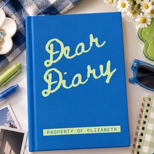 May include: A blue hardbound journal with the words Dear Diary written in large light green script on the front cover. Below the script, a light green rectangular label features the text PROPERTY OF ELIZABETH in dark blue capital letters.
