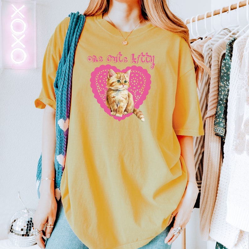 Op de afbeelding: Een geel T-shirt met een roze hart en een schattig oranje kitten erin. De tekst "one cute kitty!" staat op het shirt gedrukt.