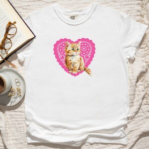 May include: White t-shirt with a pink heart-shaped doily featuring a cute orange kitten inside.
