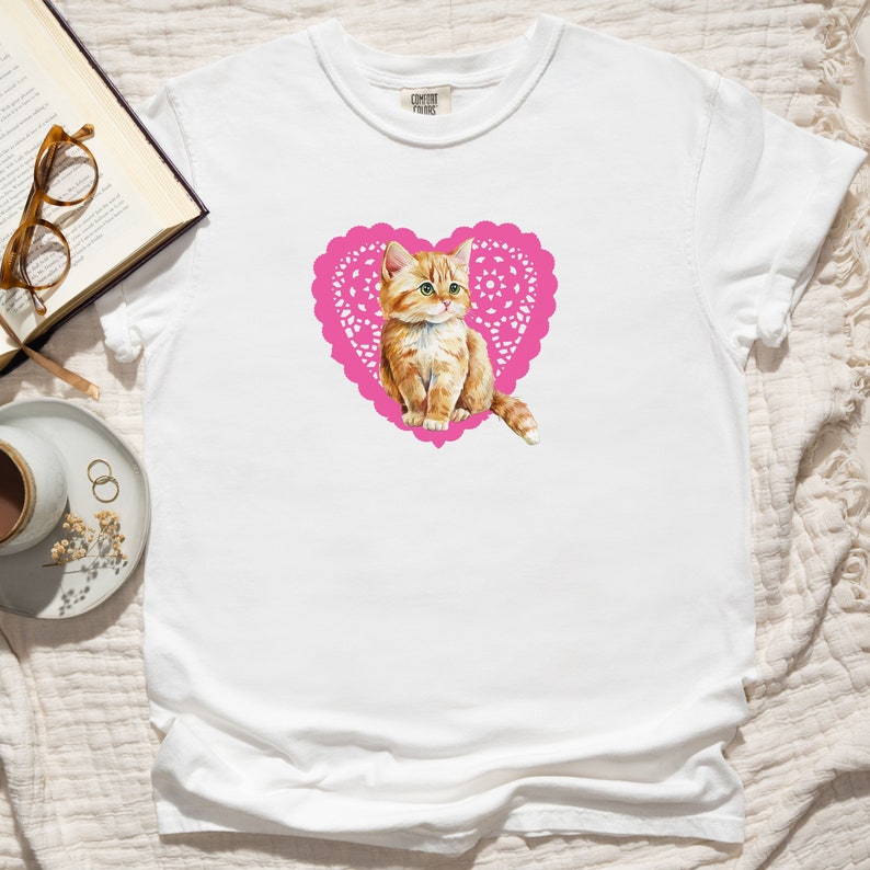 May include: White t-shirt with a pink heart-shaped doily featuring a cute orange kitten inside.