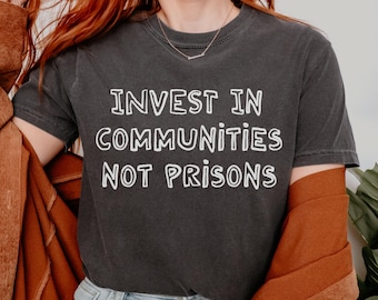May include: Dark gray t-shirt with the white text "INVEST IN COMMUNITIES NOT PRISONS." The shirt has short sleeves and a crew neck. The image shows a person wearing the shirt.