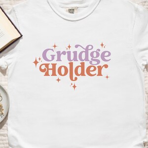 May include: White t-shirt with the text "Grudge Holder" in purple and orange lettering with small star accents.