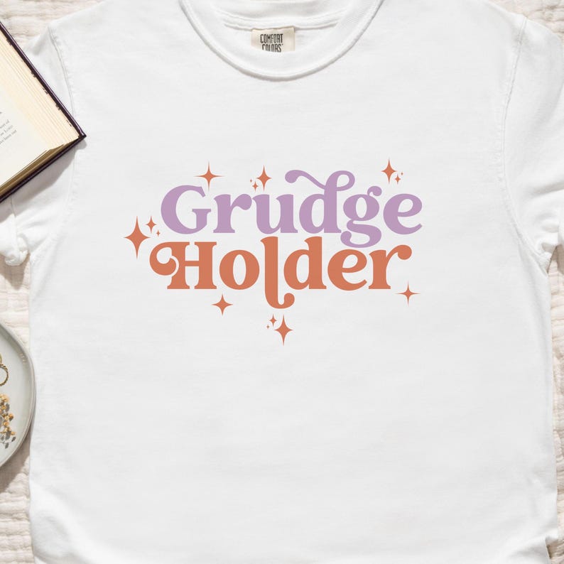 May include: White t-shirt with the text "Grudge Holder" in purple and orange lettering with small star accents.