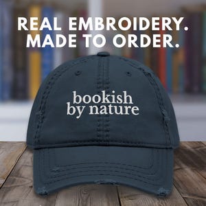 May include: A distressed navy blue baseball cap with the embroidered text "bookish by nature" in white. The cap has a curved brim and a worn appearance. The text "REAL EMBROIDERY. MADE TO ORDER." is above the cap.