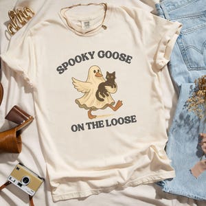 May include: Cream-colored t-shirt with the text "Spooky Goose on the Loose" and a cartoon graphic of a goose in a ghost costume holding a black cat. The shirt is styled with blue jeans, brown leather shoes, and a vintage camera.