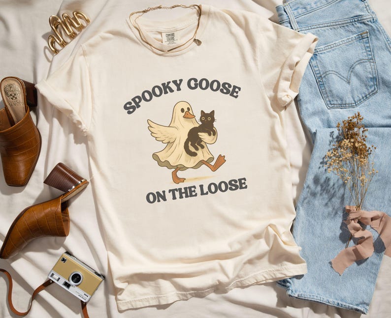 May include: Cream-colored t-shirt with the text "Spooky Goose on the Loose" and a cartoon graphic of a goose in a ghost costume holding a black cat. The shirt is styled with blue jeans, brown leather shoes, and a vintage camera.