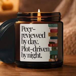 May include: A lit candle in a brown glass jar with a black lid. The label reads "Peer-reviewed by day. Plot-driven by night." A stack of colourful books is illustrated on the label. The candle sits on a wooden surface.