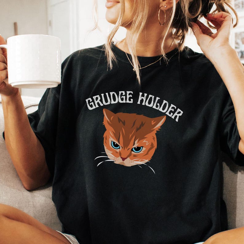 May include: Black t-shirt with a graphic of an orange cat with blue eyes and the text "GRUDGE HOLDER" in white.