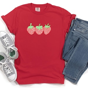 Może przedstawiać: Czerwona koszulka z nadrukiem trzech truskawek w kratkę. Truskawki są zielone i czerwone z białymi wzorami w kratkę.