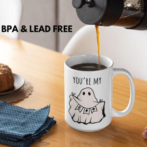 May include: A white ceramic mug with a black handle is being filled with coffee. The mug features a ghost illustration with the text "YOU'RE MY BOO". The words "BPA & LEAD FREE" are printed above the mug.