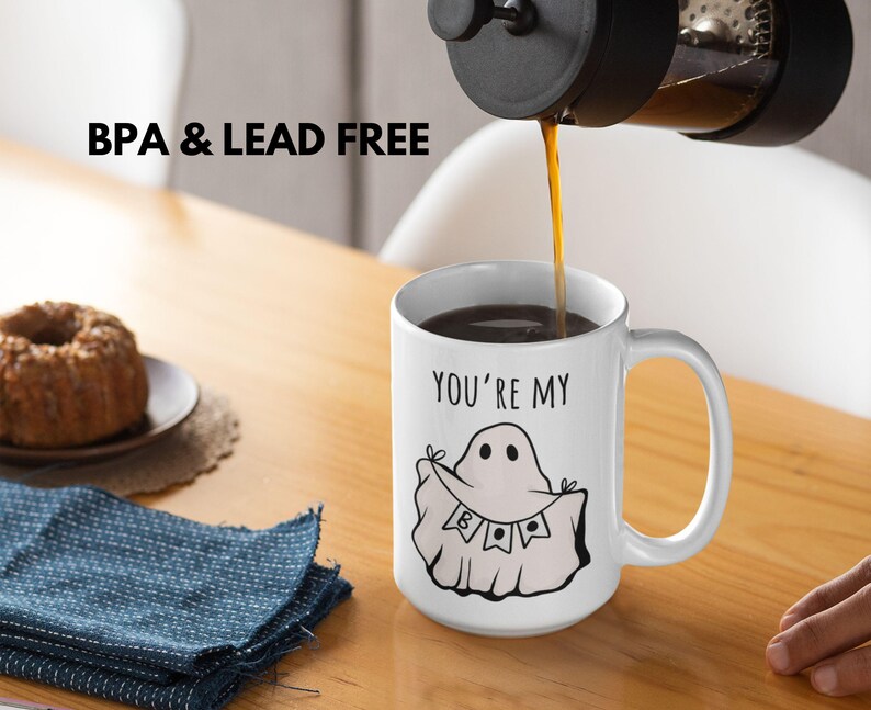 May include: A white ceramic mug with a black handle is being filled with coffee. The mug features a ghost illustration with the text "YOU'RE MY BOO". The words "BPA & LEAD FREE" are printed above the mug.
