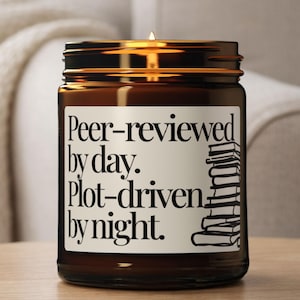 May include: A brown glass jar candle with a gold lid. The label reads "Peer-reviewed by day. Plot-driven by night." A stack of books is illustrated on the label. The candle is lit, with a small flame visible.
