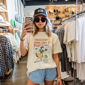 May include: A cream-coloured t-shirt featuring the text "SOME LIKE IT PLOT-DRIVEN" and a retro illustration of a woman with books. The shirt is paired with blue and white checked shorts. The photo is taken in a shop.