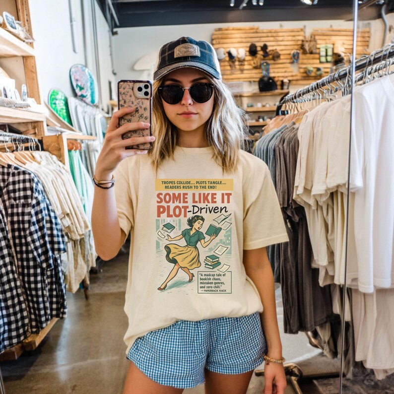 May include: A cream-coloured t-shirt featuring the text "SOME LIKE IT PLOT-DRIVEN" and a retro illustration of a woman with books. The shirt is paired with blue and white checked shorts. The photo is taken in a shop.