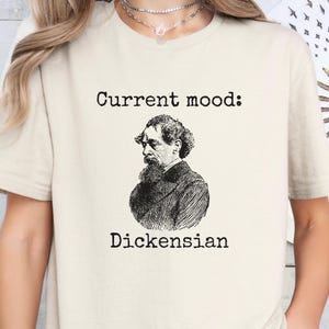 Puede incluir: Una camiseta blanca con el texto "Current mood: Dickensian" y una ilustración en blanco y negro de Charles Dickens.
